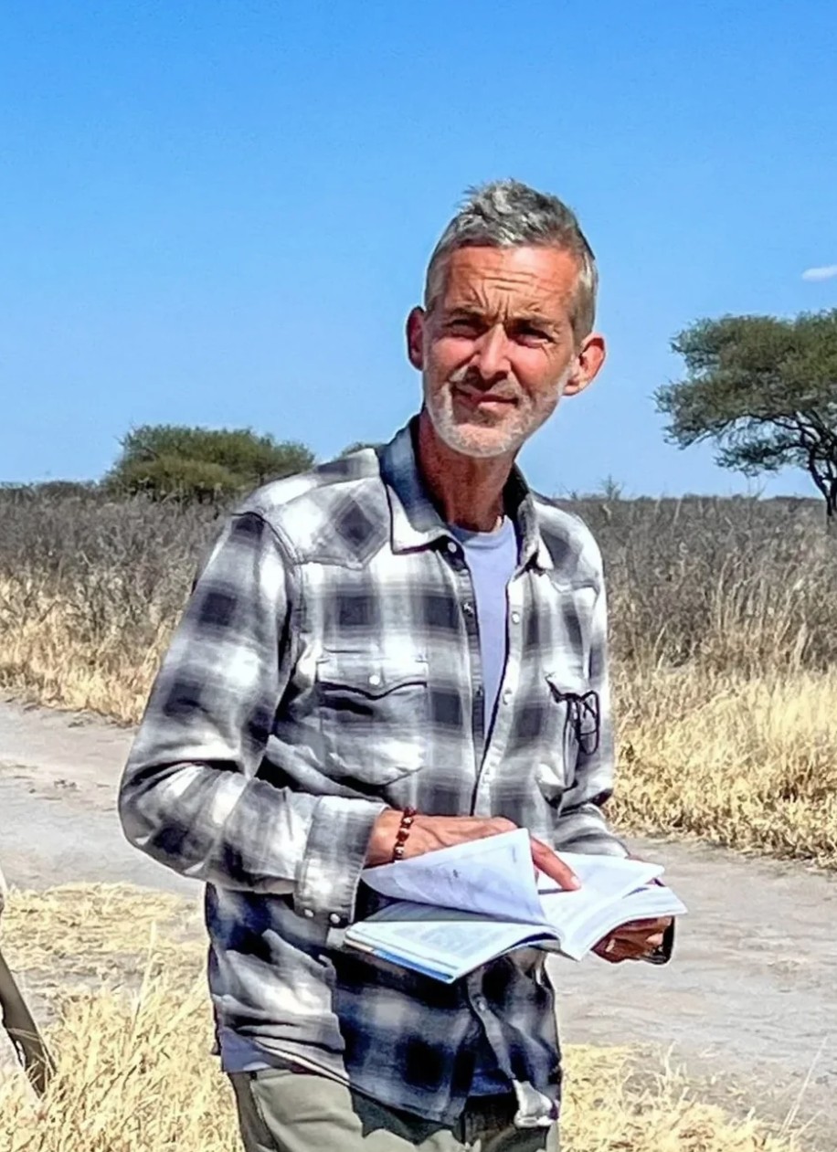 Allan Hancock College film instructor Tim Webb stands outdoors on a rural dirt road holding an open notebook. Dressed in a plaid overshirt, he looks toward the camera with dry grass, scattered trees, and open landscape behind him, reflecting a location scouting or filmmaking setting.